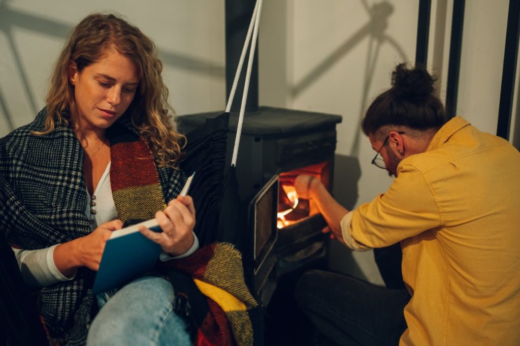 Un couple qui se réchauffe avec un poêle à bois et l'homme y ajoute des bois